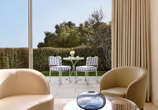 Elegant lounge area with a curved sofa, modern armchair, and glass coffee table overlooking a private terrace with striped chairs and a table set against a backdrop of greenery.