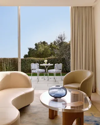 Elegant lounge area with a curved sofa, modern armchair, and glass coffee table overlooking a private terrace with striped chairs and a table set against a backdrop of greenery.