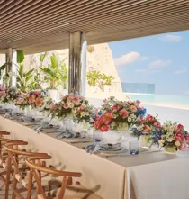 Longue table dressée avec nappes blanches, chaises en bois, compositions florales colorées et vue sur la mer, sous une terrasse ombragée.