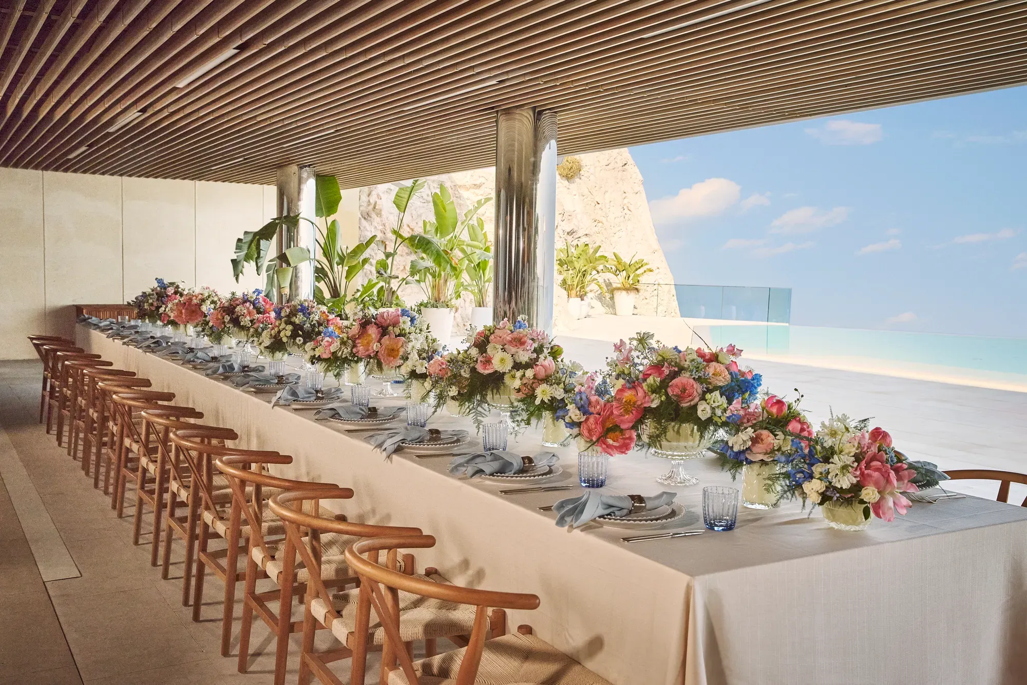 Longue table dressée avec nappes blanches, chaises en bois, compositions florales colorées et vue sur la mer, sous une terrasse ombragée.