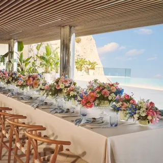 Long table set with white linens, wooden chairs, colourful floral arrangements, and sea views under a shaded terrace.