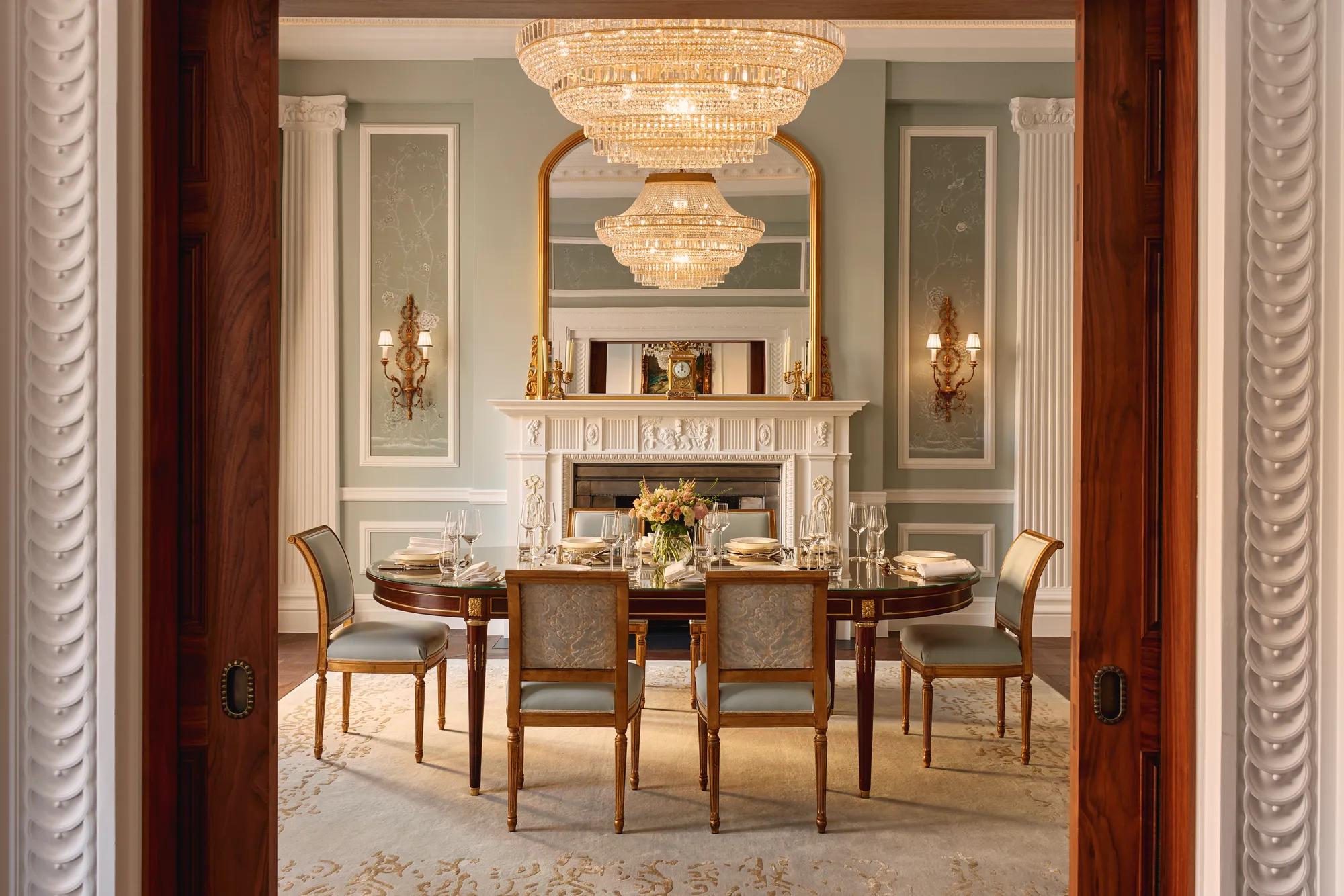 Formal dining room with an oval table set for eight, blue and gold upholstered chairs, and a grand chandelier above, framed by ornate columns and a decorative white fireplace with a gilded mirror.