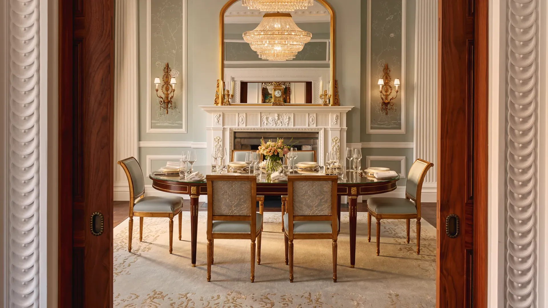 Formal dining room with an oval table set for eight, blue and gold upholstered chairs, and a grand chandelier above, framed by ornate columns and a decorative white fireplace with a gilded mirror.