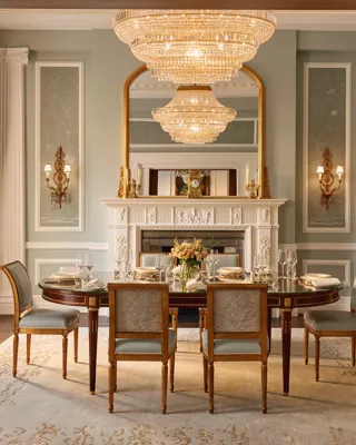 Formal dining room with an oval table set for eight, blue and gold upholstered chairs, and a grand chandelier above, framed by ornate columns and a decorative white fireplace with a gilded mirror.
