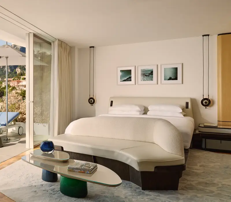 A bright suite bedroom with a terrace, mountain views, and a sun lounger outside sliding glass doors.