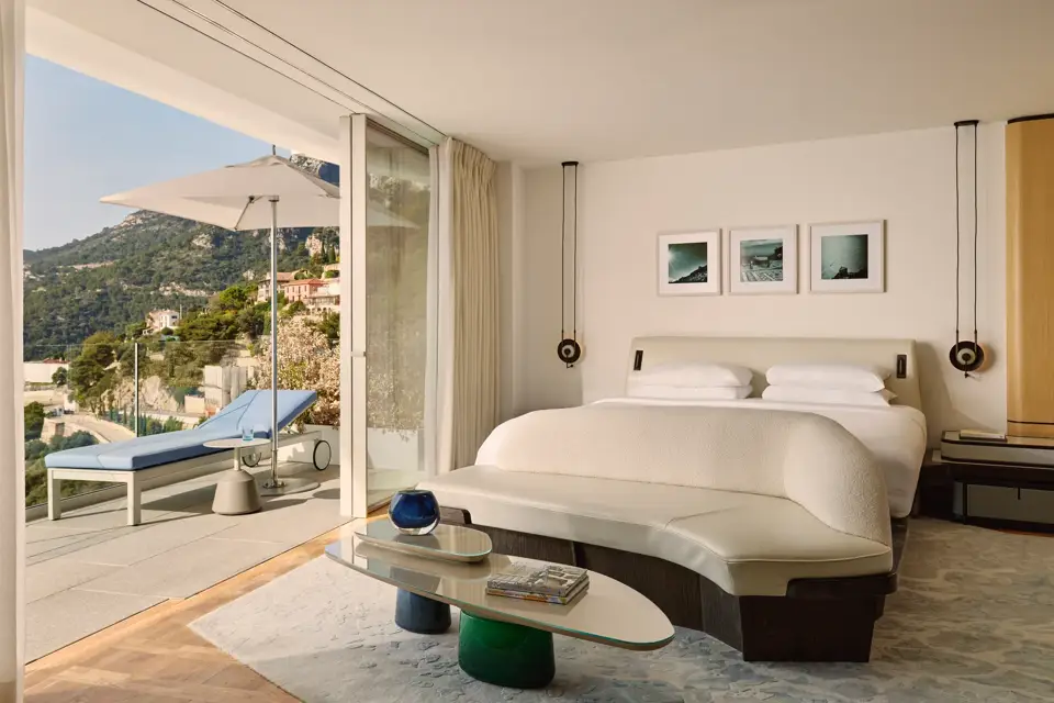 A bright suite bedroom with a terrace, mountain views, and a sun lounger outside sliding glass doors.
