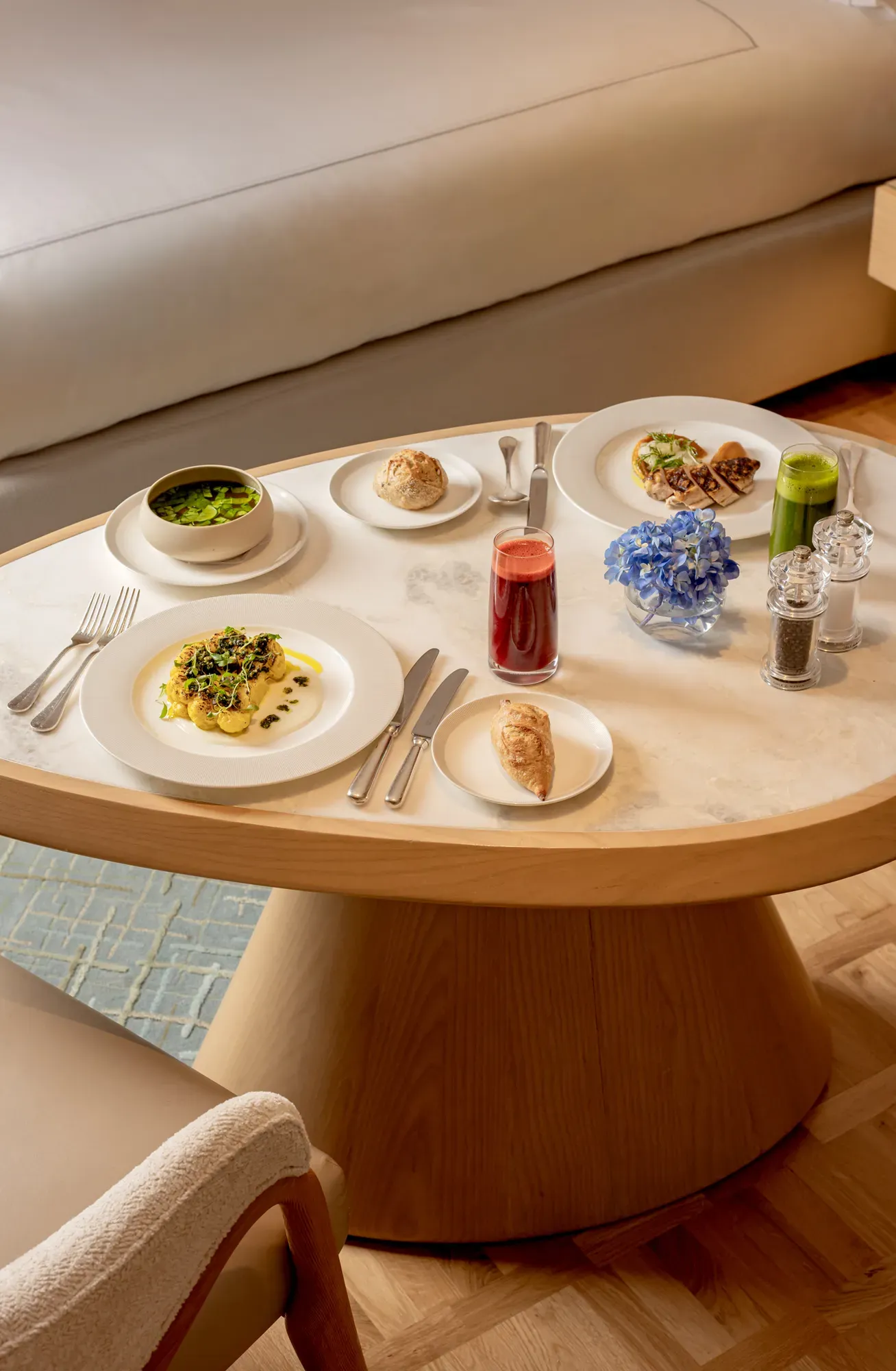 In-room dining table with plated dishes, juice, pastries and flowers beside a sofa in a bright hotel suite.