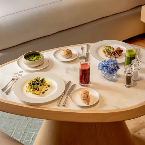 In-room dining table with plated dishes, juice, pastries and flowers beside a sofa in a bright hotel suite.