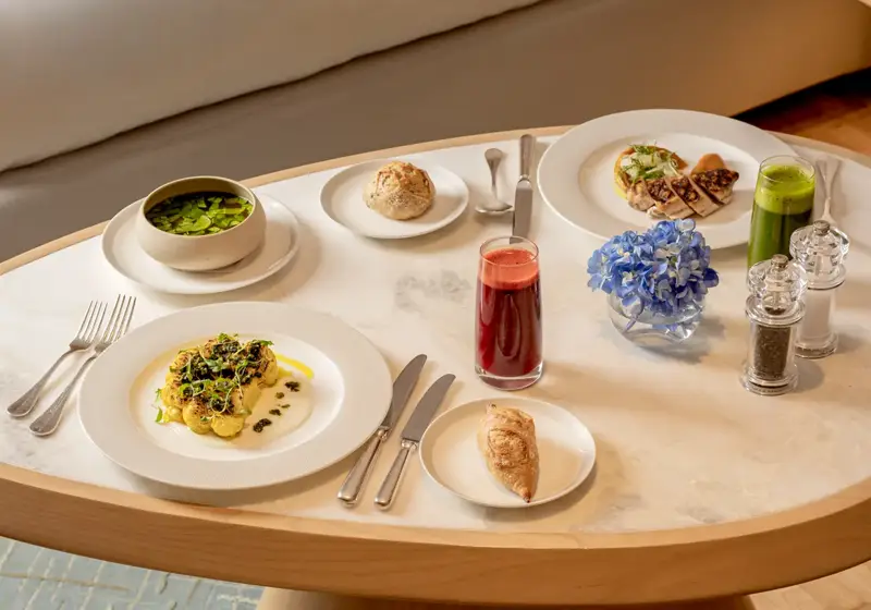 In-room dining table with plated dishes, juice, pastries and flowers beside a sofa in a bright hotel suite.