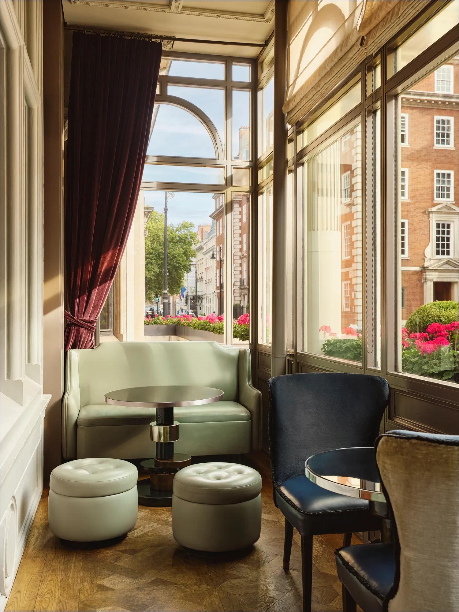 Sunlit seating nook with pastel sofa, round stools, and large windows overlooking a street with flowers and brick buildings.