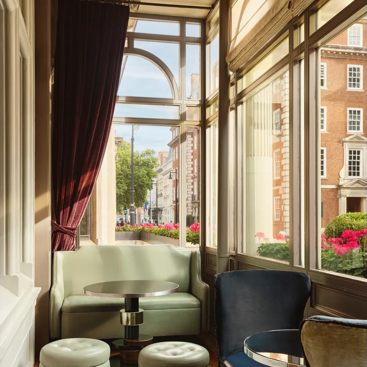 Sunlit seating nook with pastel sofa, round stools, and large windows overlooking a street with flowers and brick buildings.