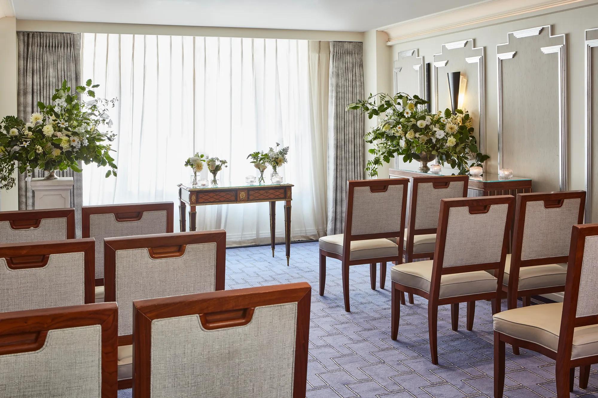 Intimate ceremony room at Claridge’s with rows of wooden chairs facing a small ornate table, surrounded by large floral arrangements and softly lit by natural light through sheer curtains.