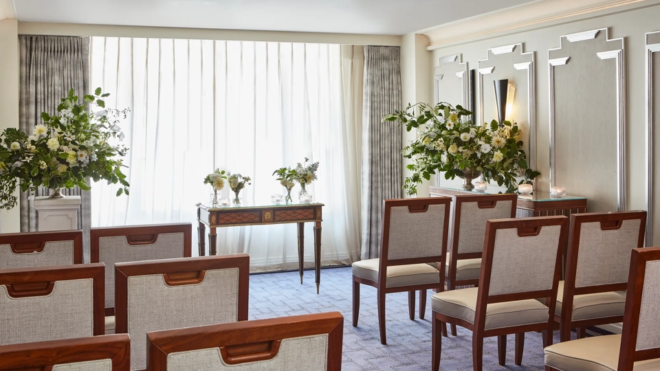 Intimate ceremony room at Claridge’s with rows of wooden chairs facing a small ornate table, surrounded by large floral arrangements and softly lit by natural light through sheer curtains.
