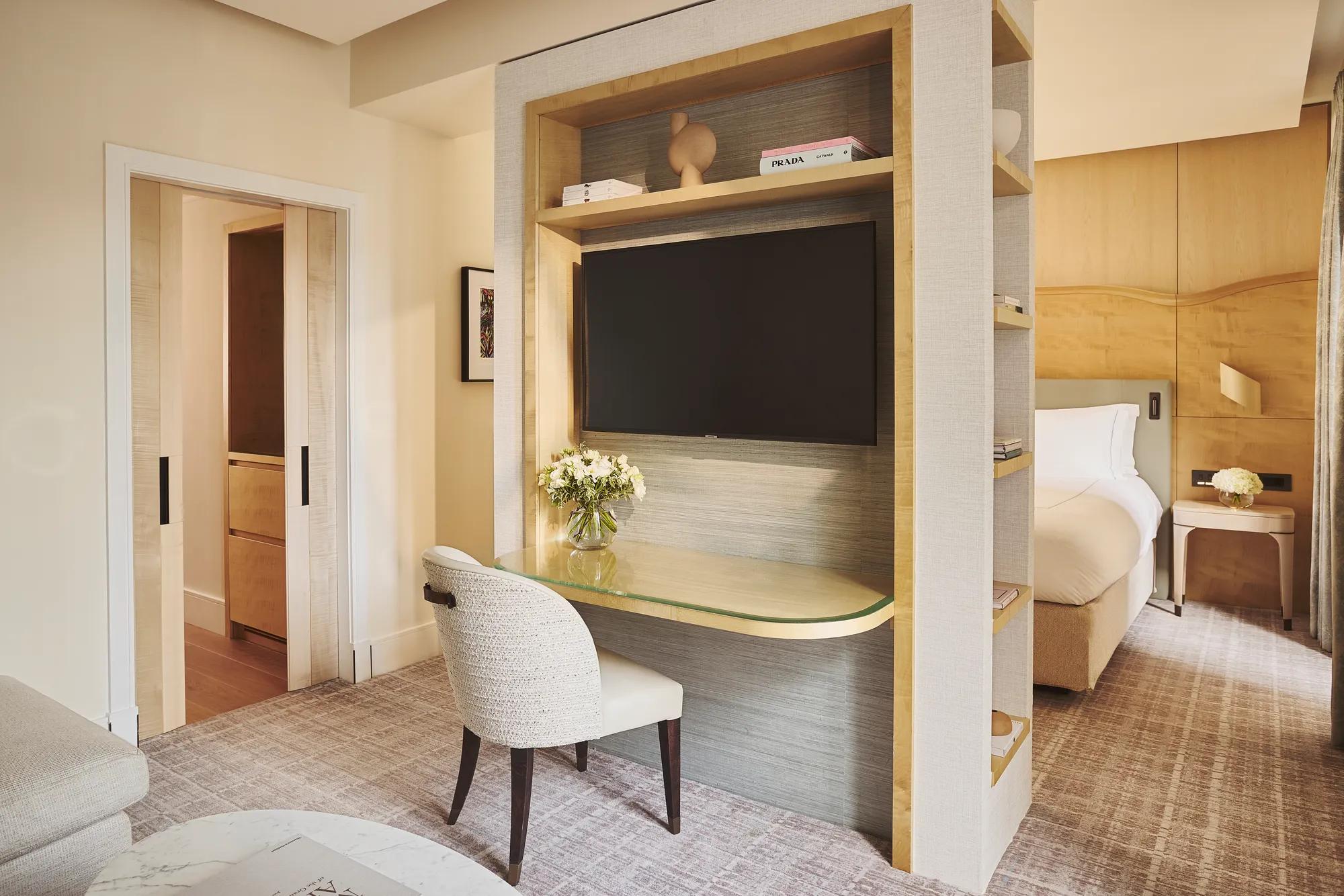 Desk with chair and built-in shelving beneath a wall-mounted TV, beside sliding doors and a bedroom with bed and bedside table beyond.
