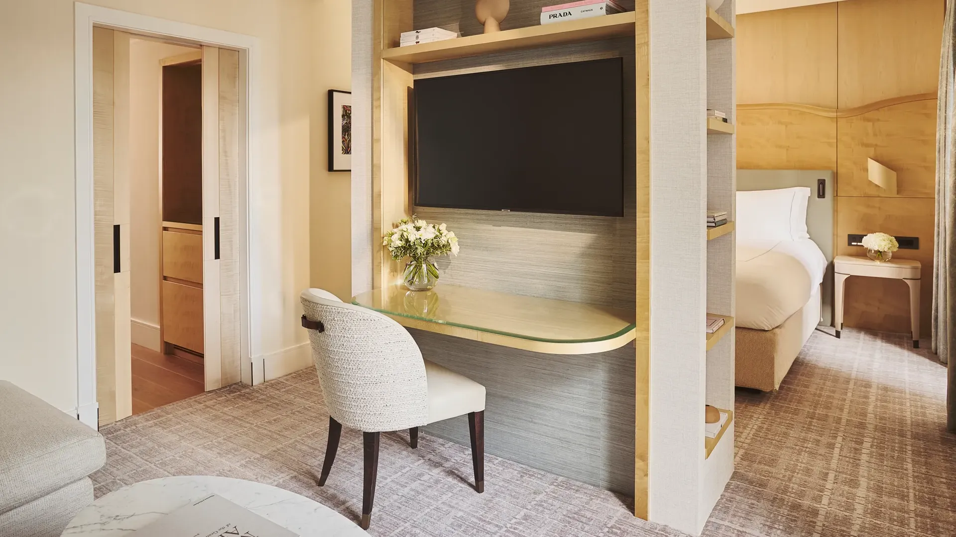 Desk with chair and built-in shelving beneath a wall-mounted TV, beside sliding doors and a bedroom with bed and bedside table beyond.