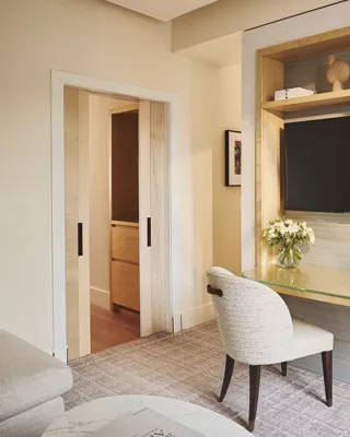 Desk with chair and built-in shelving beneath a wall-mounted TV, beside sliding doors and a bedroom with bed and bedside table beyond.