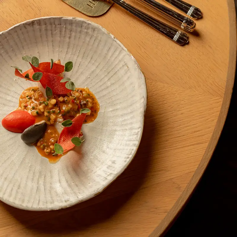 Delicately plated dish with a creamy, finely chopped mixture, red and black garnishes and micro herbs, served in a textured white bowl on a wooden table.