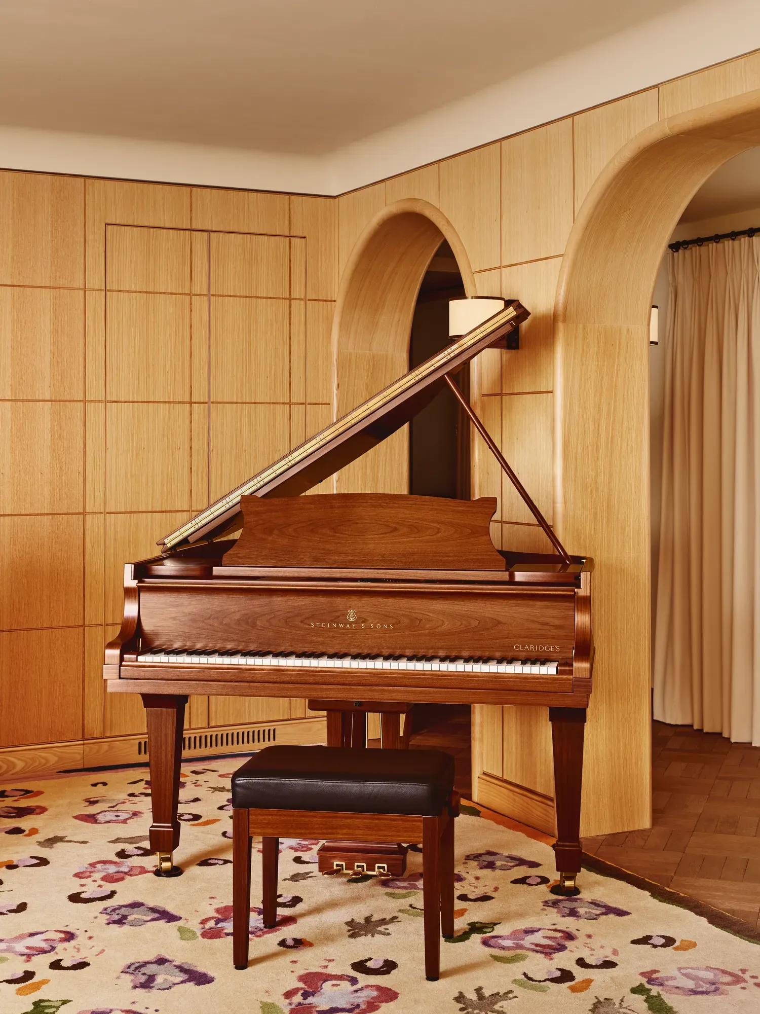 Steinway & Sons grand piano in polished walnut with a black leather stool, set against curved wooden archways and pale wood wall panelling on a floral-patterned rug.