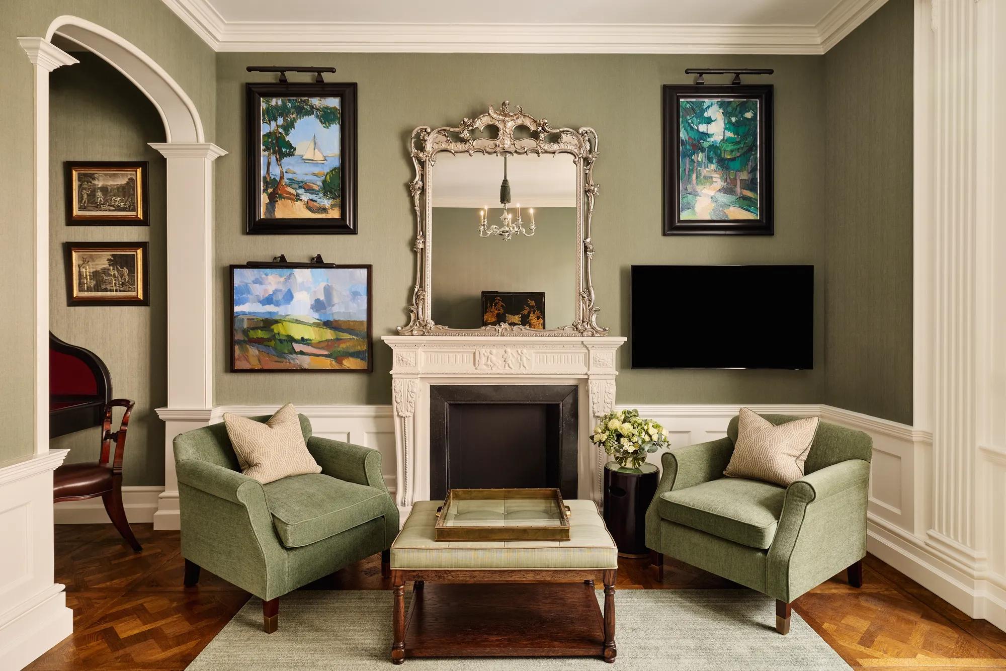 Elegant sitting room with two green armchairs, ornate mirror above a white fireplace, and framed artwork on the walls.