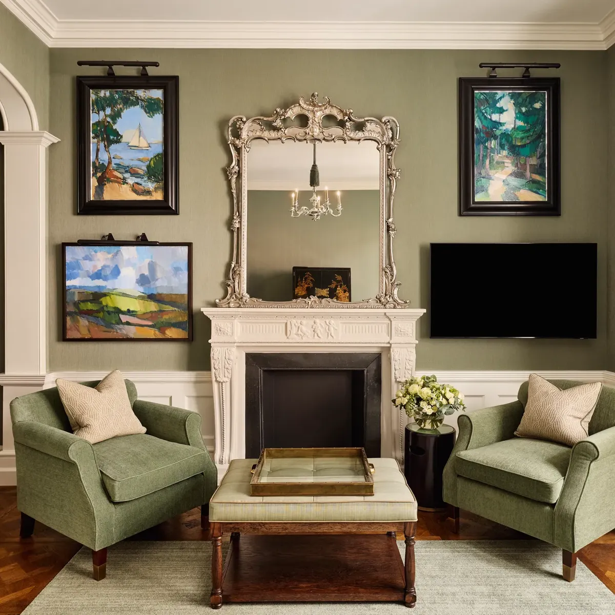 Elegant sitting room with two green armchairs, ornate mirror above a white fireplace, and framed artwork on the walls.