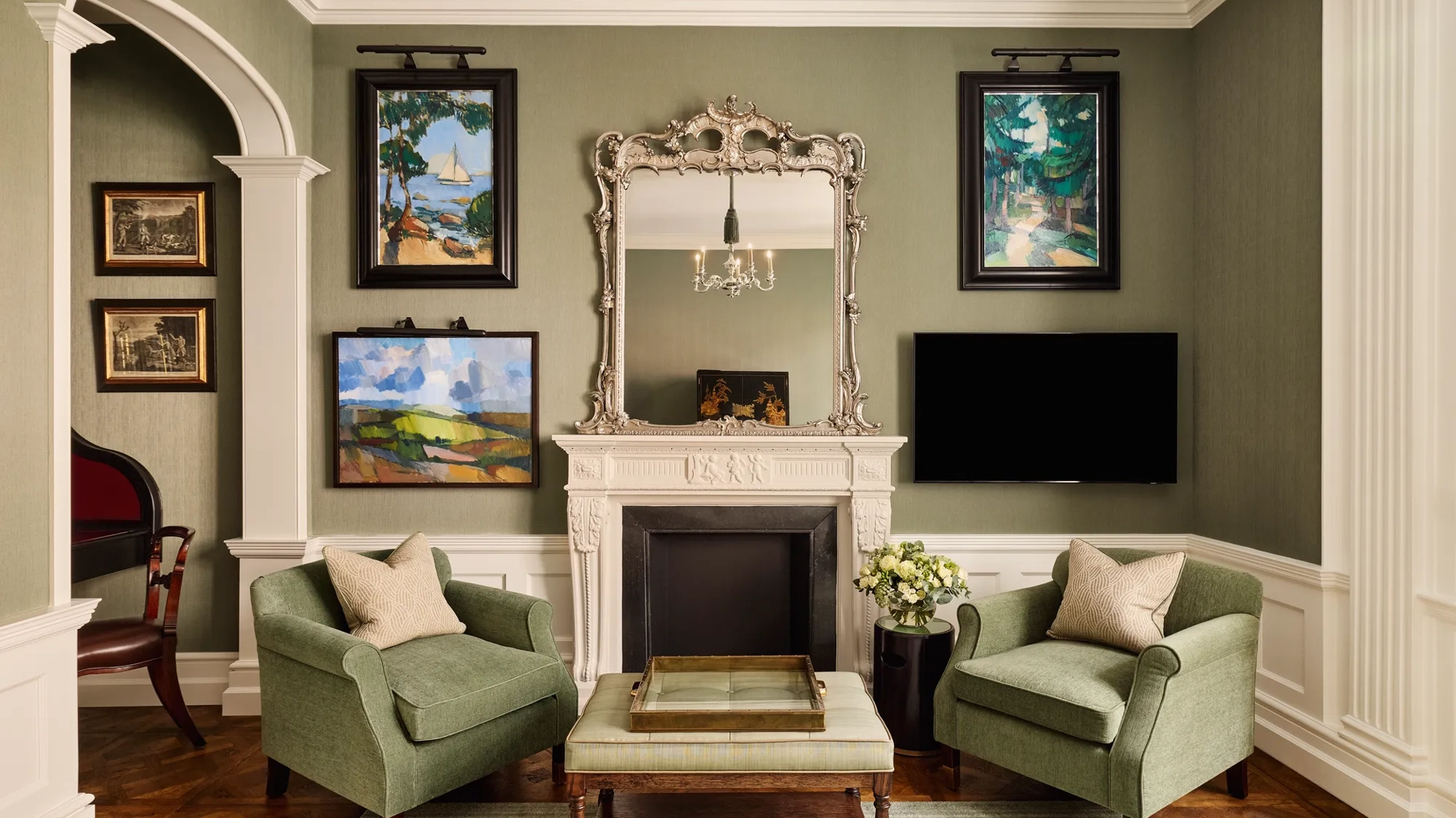 Elegant sitting room with two green armchairs, ornate mirror above a white fireplace, and framed artwork on the walls.