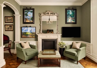 Elegant sitting room with two green armchairs, ornate mirror above a white fireplace, and framed artwork on the walls.