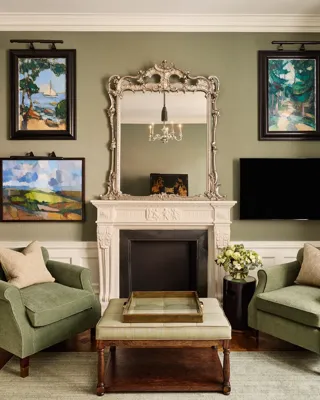 Elegant sitting room with two green armchairs, ornate mirror above a white fireplace, and framed artwork on the walls.
