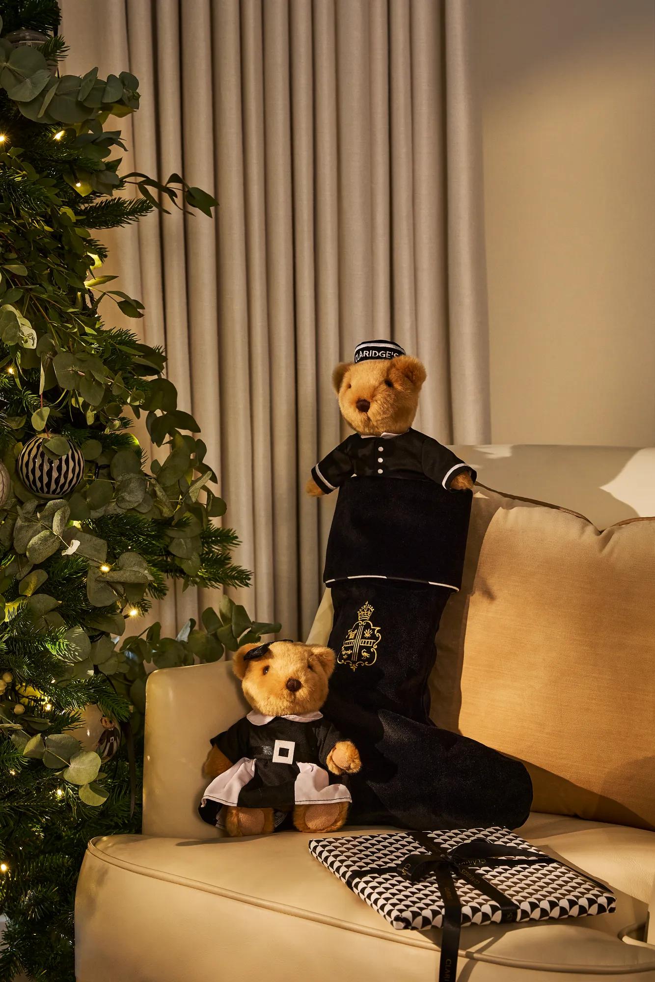 Claridge’s teddy bears beside a Christmas stocking and wrapped gift by a decorated tree.