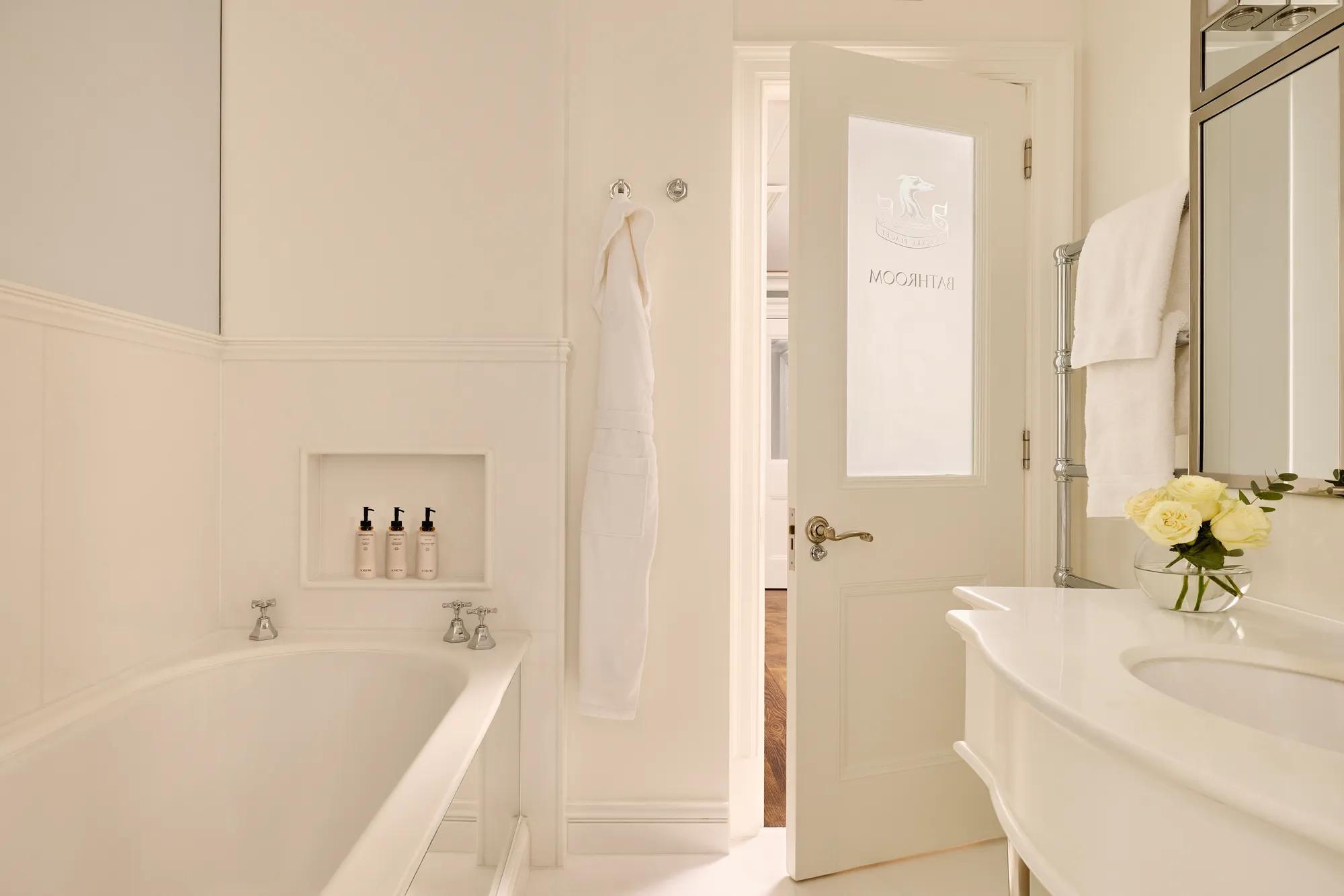Light, elegant bathroom with a bathtub, white robes, marble sink, and a door etched with “Bathroom.”