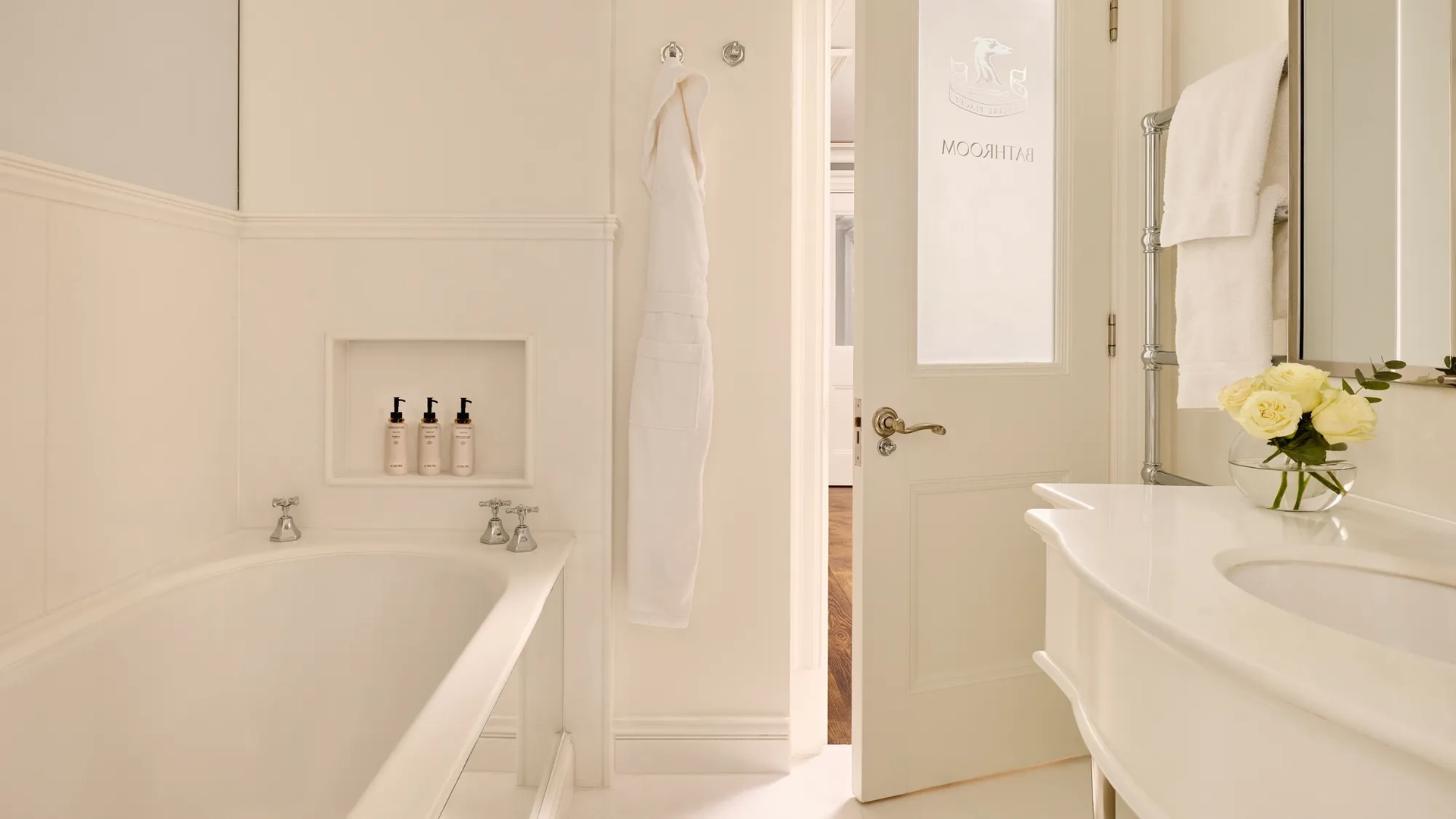 Light, elegant bathroom with a bathtub, white robes, marble sink, and a door etched with “Bathroom.”