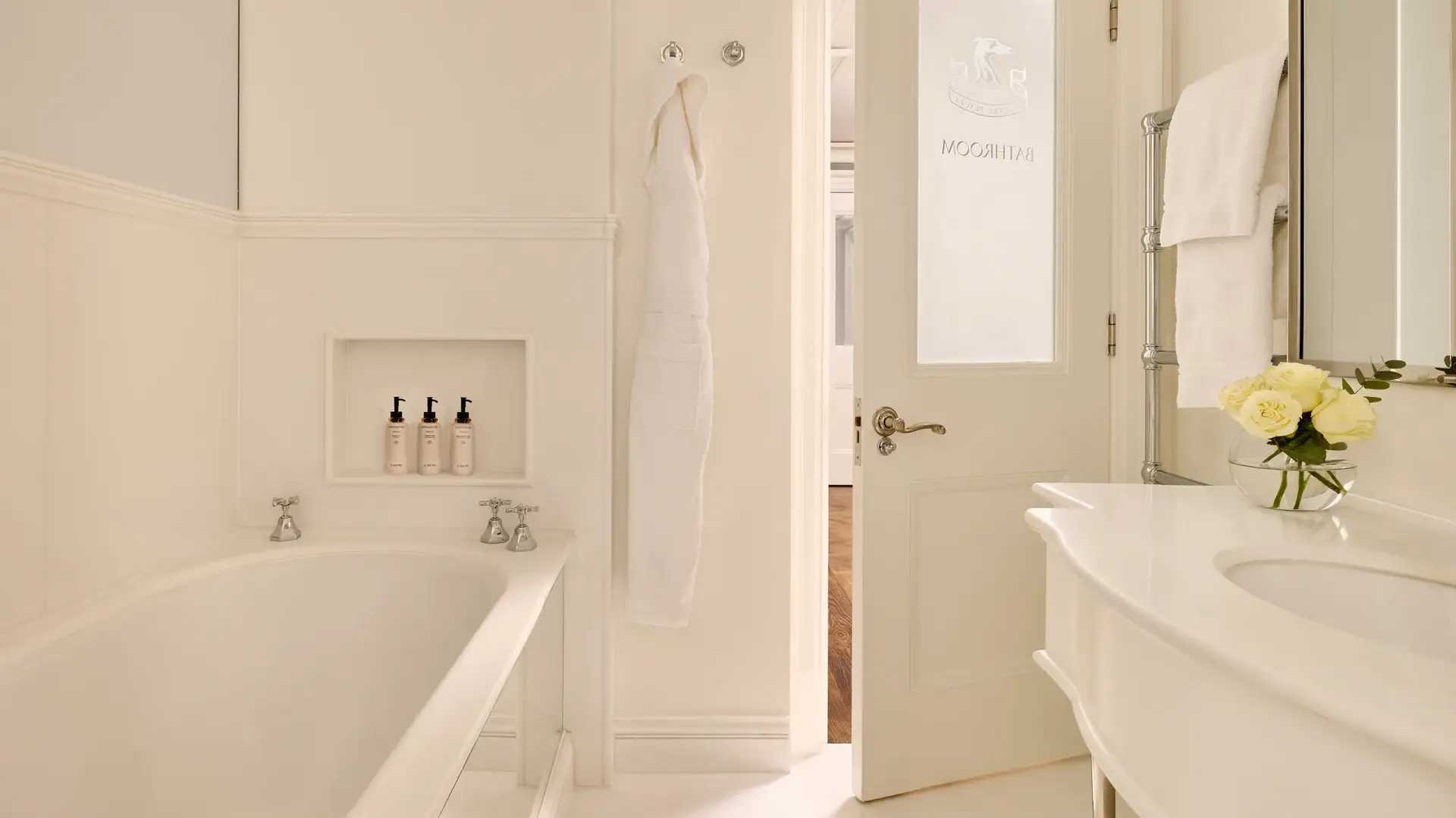 Light, elegant bathroom with a bathtub, white robes, marble sink, and a door etched with “Bathroom.”