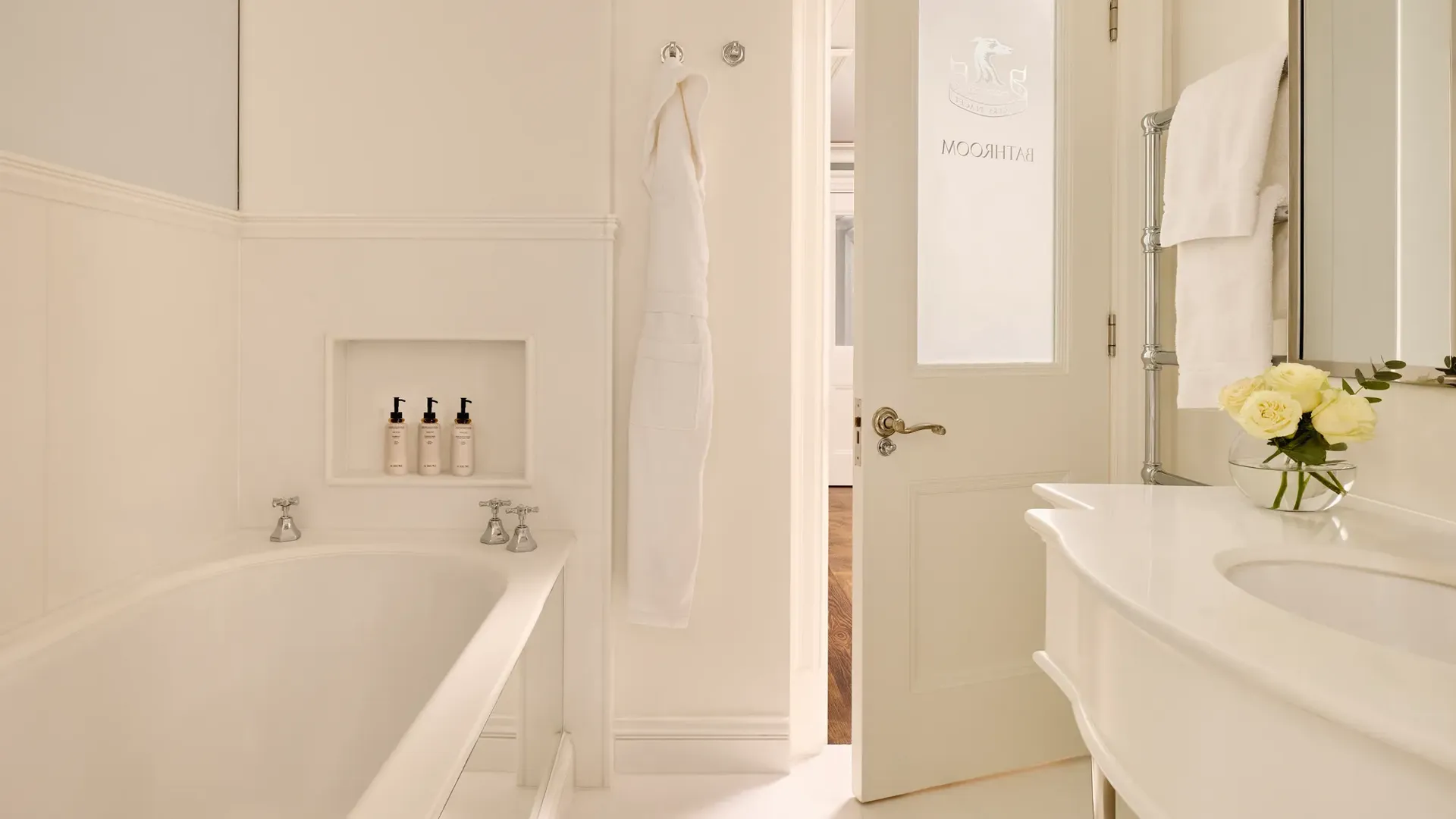 Light, elegant bathroom with a bathtub, white robes, marble sink, and a door etched with “Bathroom.”
