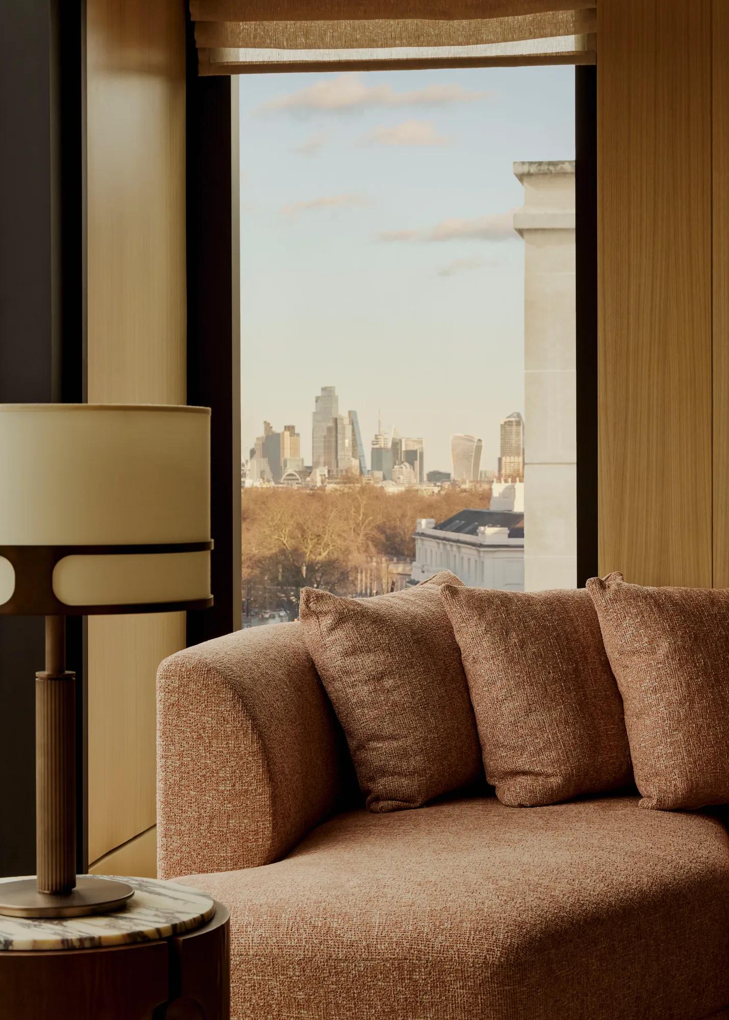 Upholstered sofa beside a floor-to-ceiling window with side table and lamp, overlooking treetops and a distant city skyline.