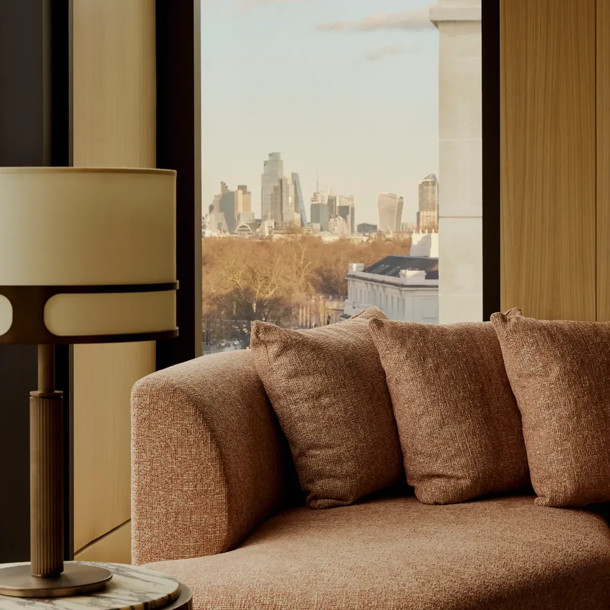 Upholstered sofa beside a floor-to-ceiling window with side table and lamp, overlooking treetops and a distant city skyline.