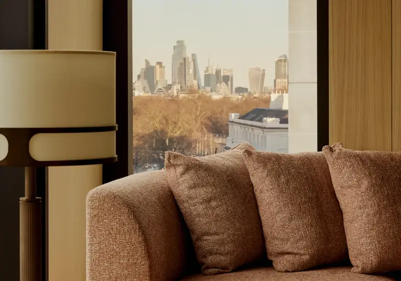 Upholstered sofa beside a floor-to-ceiling window with side table and lamp, overlooking treetops and a distant city skyline.