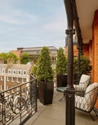 Balcony with wrought-iron railings and potted greenery overlooking red-brick and stone buildings, featuring two cushioned chairs and a small round table set against the backdrop of a clear blue sky.