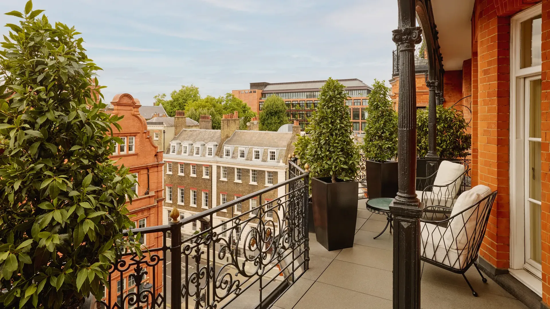 Balcony with wrought-iron railings and potted greenery overlooking red-brick and stone buildings, featuring two cushioned chairs and a small round table set against the backdrop of a clear blue sky.
