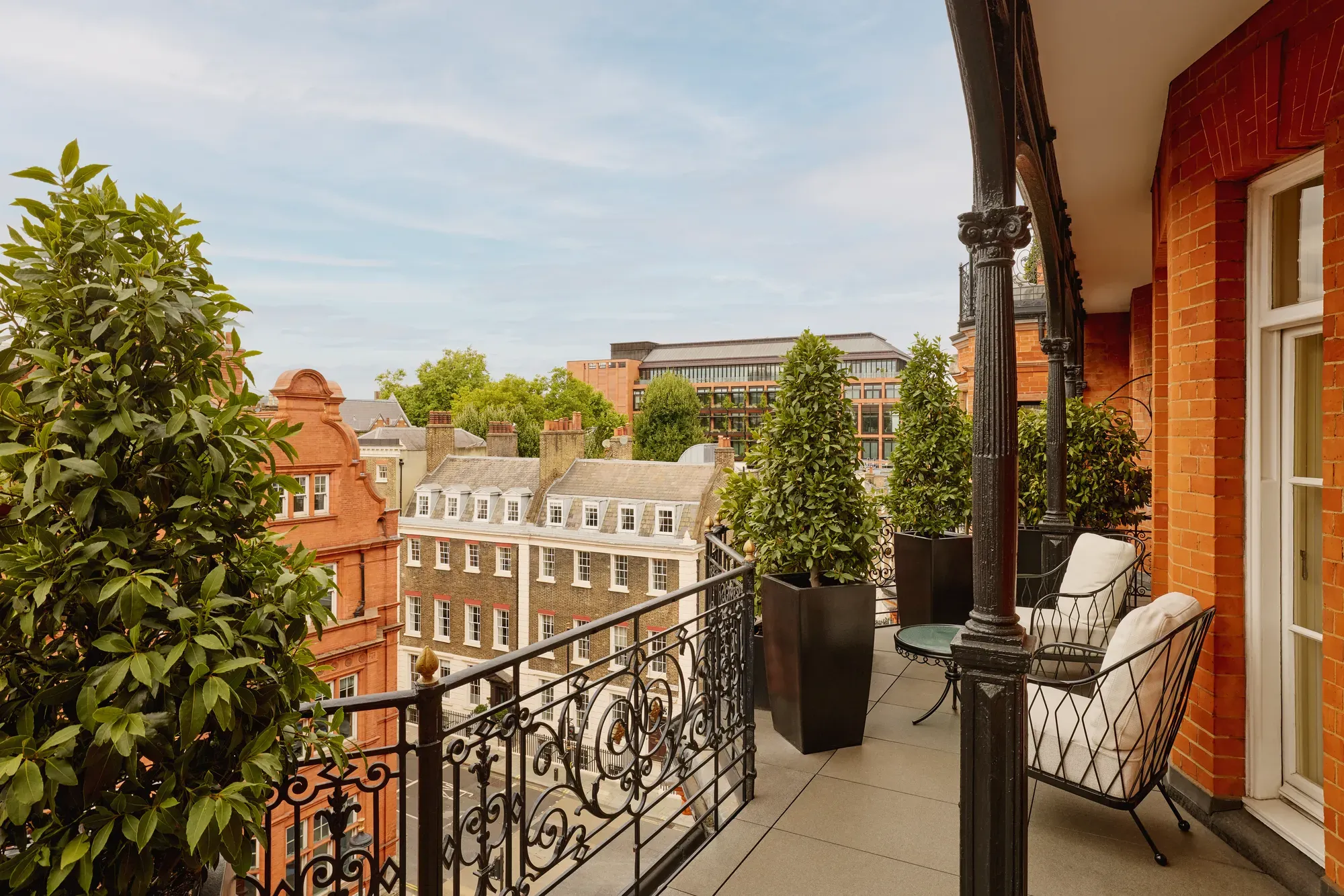 Balcony with wrought-iron railings and potted greenery overlooking red-brick and stone buildings, featuring two cushioned chairs and a small round table set against the backdrop of a clear blue sky.