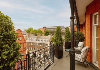 Balcony with wrought-iron railings and potted greenery overlooking red-brick and stone buildings, featuring two cushioned chairs and a small round table set against the backdrop of a clear blue sky.