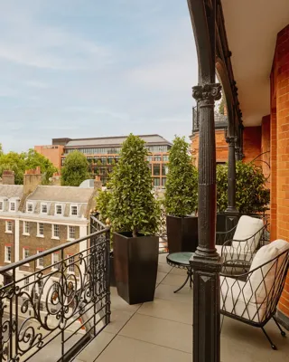 Balcony with wrought-iron railings and potted greenery overlooking red-brick and stone buildings, featuring two cushioned chairs and a small round table set against the backdrop of a clear blue sky.
