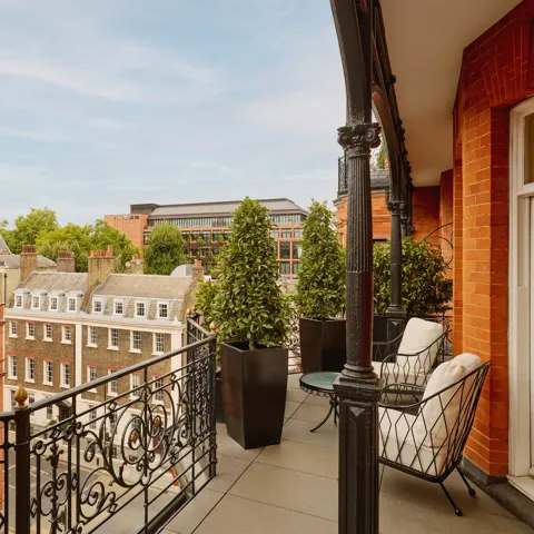 Balcony with wrought-iron railings and potted greenery overlooking red-brick and stone buildings, featuring two cushioned chairs and a small round table set against the backdrop of a clear blue sky.