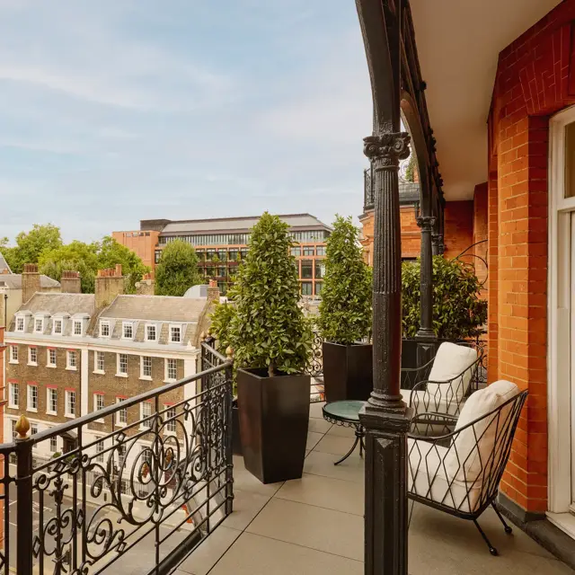 Balcony with wrought-iron railings and potted greenery overlooking red-brick and stone buildings, featuring two cushioned chairs and a small round table set against the backdrop of a clear blue sky.