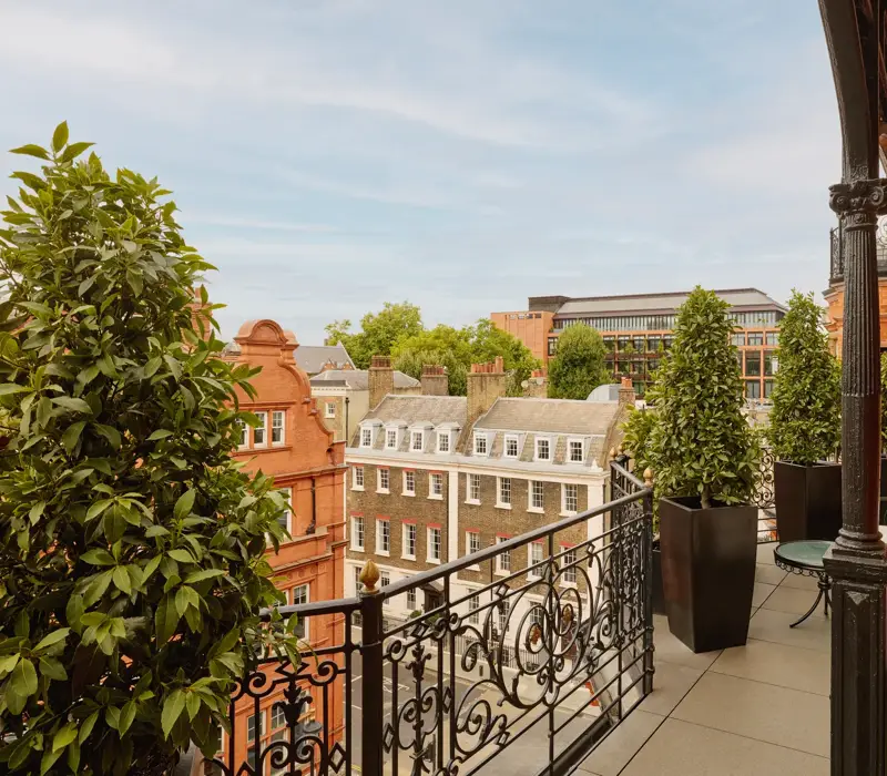 Balcony with wrought-iron railings and potted greenery overlooking red-brick and stone buildings, featuring two cushioned chairs and a small round table set against the backdrop of a clear blue sky.