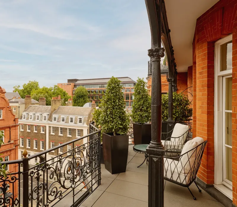 Balcony with wrought-iron railings and potted greenery overlooking red-brick and stone buildings, featuring two cushioned chairs and a small round table set against the backdrop of a clear blue sky.