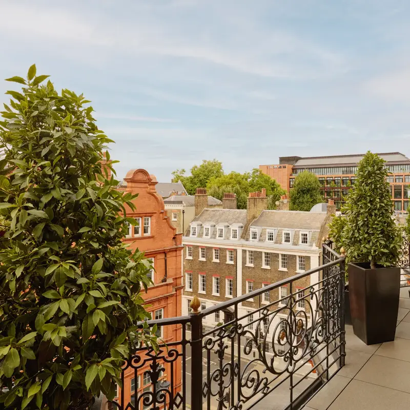 Balcony with wrought-iron railings and potted greenery overlooking red-brick and stone buildings, featuring two cushioned chairs and a small round table set against the backdrop of a clear blue sky.