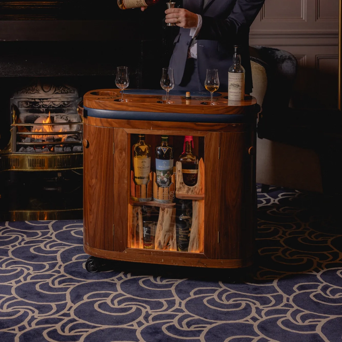 Server in a dark suit pouring whisky at a wooden drinks trolley in front of a lit fireplace.