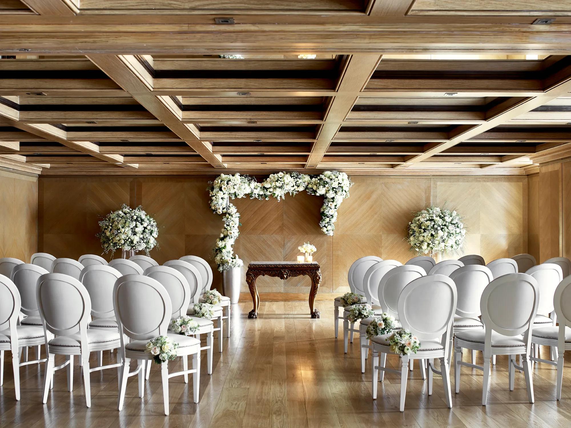 Wedding ceremony room with rows of white chairs facing a floral arch and ornate wooden table under a coffered ceiling.