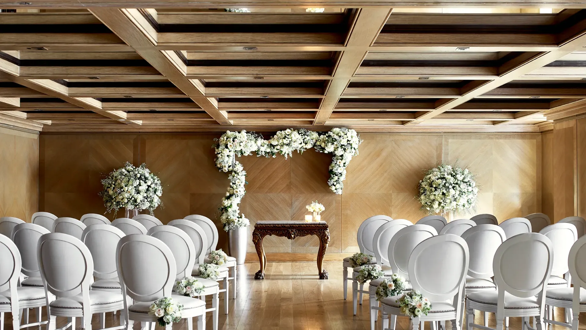Wedding ceremony room with rows of white chairs facing a floral arch and ornate wooden table under a coffered ceiling.