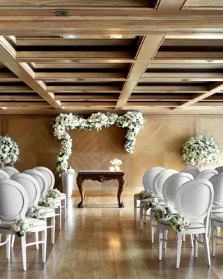 Wedding ceremony room with rows of white chairs facing a floral arch and ornate wooden table under a coffered ceiling.
