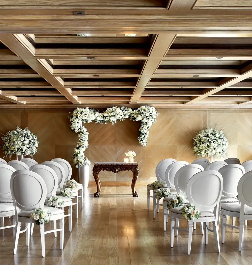 Wedding ceremony room with rows of white chairs facing a floral arch and ornate wooden table under a coffered ceiling.